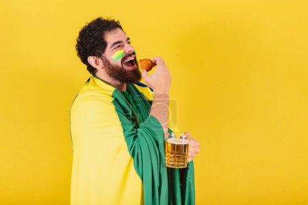 Foto de Hombre caucásico con barba, brasileño, fanático del fútbol de Brasil, sosteniendo un vaso de cerveza y un muslo de pollo, comida típica brasileña. - Imagen libre de derechos