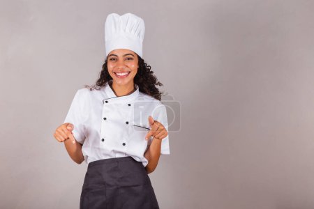 Téléchargez les photos : Jeune femme brésilienne noire, cuisinière, chef cuisinier. pointant vers le centre, seulement ici - en image libre de droit