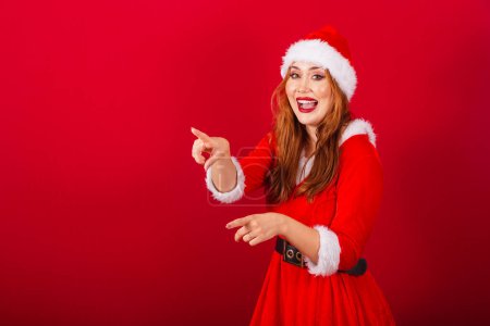 Photo for Beautiful Brazilian red-haired woman, dressed in Christmas clothes, Santa Claus. pointing at something in the distance. - Royalty Free Image