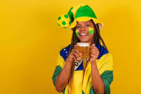 Foto de Mujer negra joven aficionada al fútbol brasileño. beber cerveza y celebrar. - Imagen libre de derechos