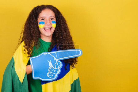 Photo for Brazilian, Caucasian, football fan girl with foam hand pointing to negative space, advertising, propaganda. and promotion. - Royalty Free Image