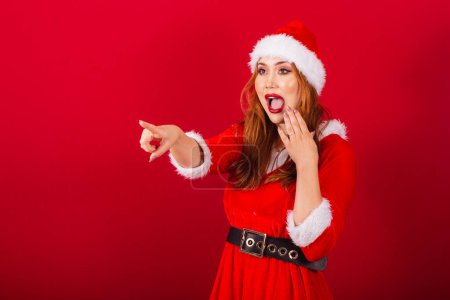 Foto de Hermosa brasileña pelirroja, vestida con ropa de Navidad, Santa Claus. apuntando a algo en la distancia. - Imagen libre de derechos