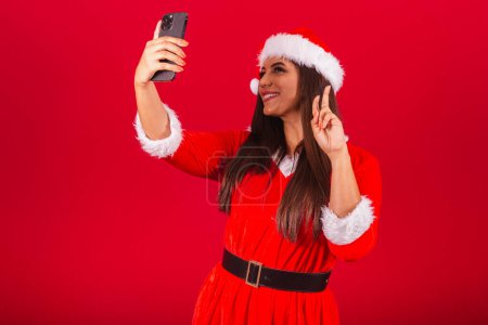 Photo for Beautiful Brazilian woman dressed in Christmas clothes, Santa Claus. holding smartphone taking self portrait. - Royalty Free Image