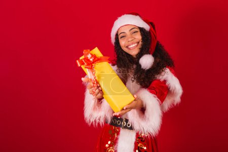 Photo for Beautiful brazilian black woman, dressed as santa claus, christmas clothes. gifting someone with yellow gift. - Royalty Free Image
