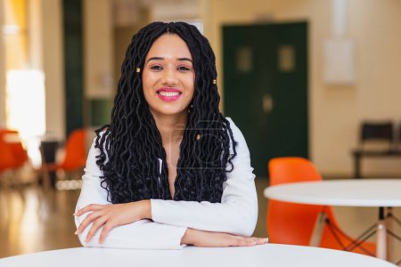 Téléchargez les photos : Jeune femme noire, brésilienne, étudiante dans un couloir universitaire souriante, vêtue de blanc. assis avec les bras croisés. - en image libre de droit