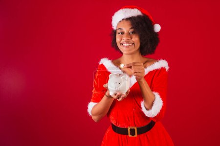 Téléchargez les photos : Belle femme brésilienne noire, habillée en Père Noël, maman Claude, tenant tirelire et pièce de monnaie - en image libre de droit
