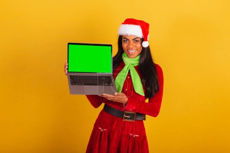 Téléchargez les photos : Belle femme brésilienne noire, vêtue de vêtements de Noël, Père Noël claus, carnet et montrant écran chromé vert. - en image libre de droit