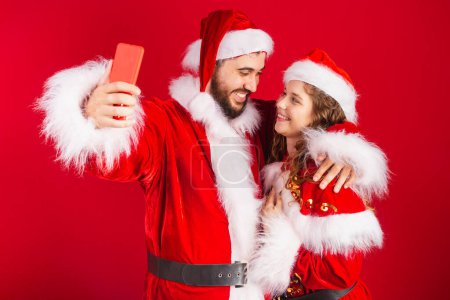 Photo for Brazilian couple, dressed in Christmas clothes, Santa Claus, using smartphone, online. - Royalty Free Image
