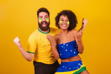 Foto de Pareja de aficionados brasileños al fútbol, vestidos con los colores de Brasil, mujer negra, hombre caucásico. Celebrando, deseando suerte al equipo - Imagen libre de derechos