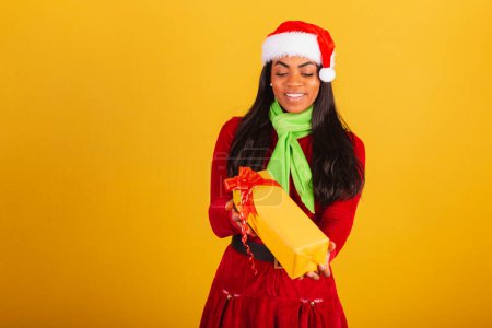 Photo for Beautiful black Brazilian woman, dressed in Christmas clothes, Santa Claus, handing yellow gift. - Royalty Free Image