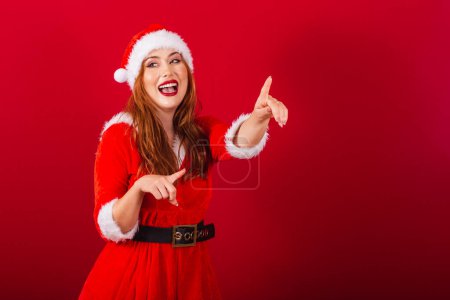 Téléchargez les photos : Belle femme rousse brésilienne, vêtue de vêtements de Noël, le Père Noël. pointant quelque chose au loin. - en image libre de droit