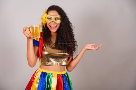 Foto de Hermosa mujer brasileña negra, con ropa frevo, carnaval. con una máscara, bailando con un vaso de cerveza, festejando en un bar. - Imagen libre de derechos