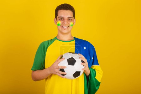 Foto de Joven aficionado al fútbol brasileño. vestido de verde, sosteniendo pelota de fútbol con bandera de Brasil. - Imagen libre de derechos