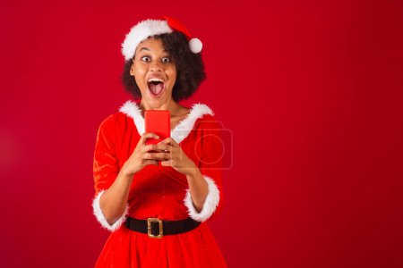 Photo for Beautiful black brazilian woman, dressed as santa claus, mama claus, watching something on smartphone - Royalty Free Image