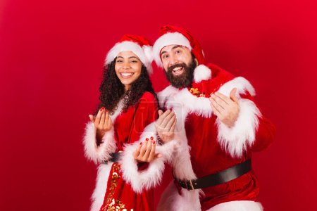 Photo for Brazilian couple, dressed for christmas, santa claus, mummy claus. calling with hands, inviting, black woman and caucasian man. - Royalty Free Image