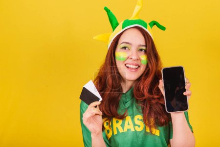 Photo for Caucasian redhead woman brazil soccer fan holding credit cards and cellphone, mobile sales concept, smartphone, app purchases. - Royalty Free Image