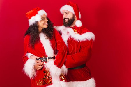 Photo for Brazilian couple, dressed for christmas, santa claus, mummy claus. black woman and caucasian man. looking at each other. - Royalty Free Image