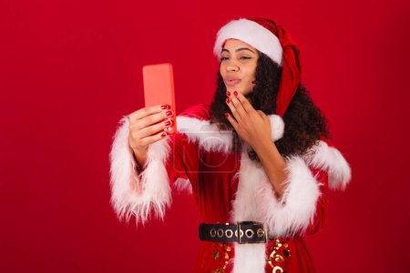 Photo for Beautiful brazilian black woman, dressed as santa claus, christmas clothes. taking self portrait by smartphone. - Royalty Free Image