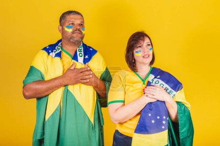 Foto de Pareja, mujer pelirroja y hombre negro, hinchas del fútbol brasileño. cantando himno nacional, patriotismo. - Imagen libre de derechos