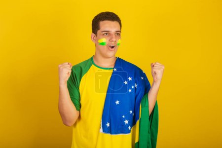 Photo for Young Brazilian man soccer fan. dressed in green, screaming goal with brazil flag. - Royalty Free Image