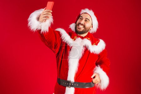 Photo for Brazilian man, dressed in santa claus clothes, holding smartphone self portrait with cellphone, video call. - Royalty Free Image