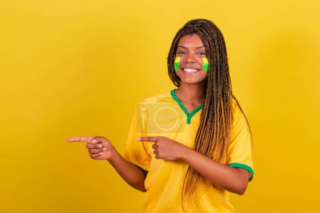 Téléchargez les photos : Femme noire jeune fan de football brésilien. pointant à gauche, souriant, publicité photo. - en image libre de droit