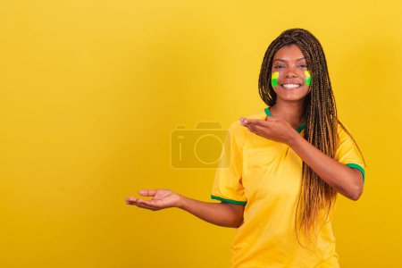 Foto de Mujer negra joven aficionada al fútbol brasileño. presentando algo a la izquierda, sonriente, foto publicitaria. - Imagen libre de derechos