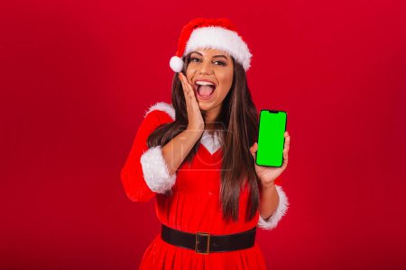 Photo for Beautiful Brazilian woman dressed in Christmas clothes, Santa Claus. holding smartphone showing green chroma screen. - Royalty Free Image