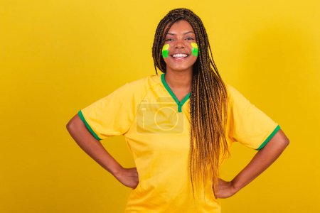 Foto de Mujer negra joven aficionada al fútbol brasileño. con las manos en las caderas, sonriendo con confianza. - Imagen libre de derechos