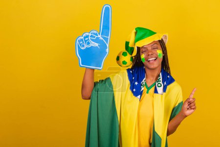 Foto de Mujer negra joven aficionada al fútbol brasileño. usando sombrero y bandera y dedo de espuma, animando campeonato. - Imagen libre de derechos
