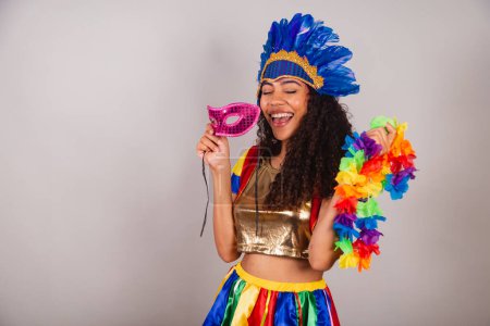 Foto de Hermosa mujer brasileña negra, con ropa frevo, carnaval. en tocado de plumas de carnaval. con máscara de carnaval y collar en las manos. - Imagen libre de derechos