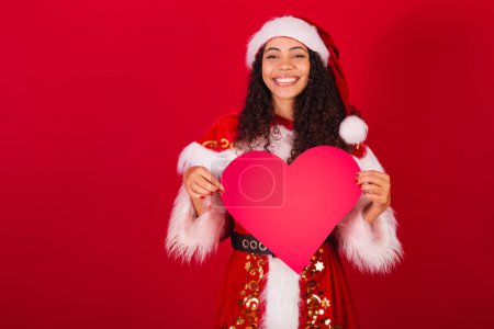 Téléchargez les photos : Belle femme noire brésilienne, habillée en Père Noël claus, vêtements de Noël. tenant coeur rouge. - en image libre de droit