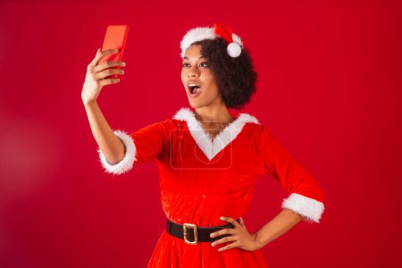 Téléchargez les photos : Belle femme brésilienne noire, habillée en Père Noël claus, maman claus, prenant autoportrait avec smartphone - en image libre de droit