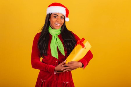 Photo for Beautiful black Brazilian woman, dressed in Christmas clothes, Santa Claus, hugging yellow gift. - Royalty Free Image