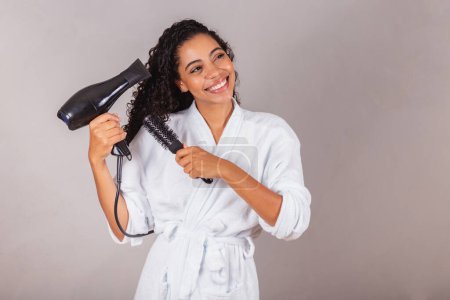 Téléchargez les photos : Belle femme noire brésilienne, portant peignoir et serviette. sèche-cheveux avec sèche-cheveux et brosse, spa, centre de beauté, soins des cheveux. - en image libre de droit