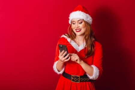 Téléchargez les photos : Belle femme rousse brésilienne, vêtue de vêtements de Noël, le Père Noël. tenant smartphone, tapant. - en image libre de droit