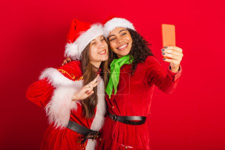 Photo for Brazilian female friends, with Christmas clothes, Santa Claus. peace and love position, selfie pose. smartphone. - Royalty Free Image