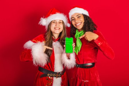 Téléchargez les photos : Des amies brésiliennes, avec des vêtements de Noël, le Père Noël. écran montrant smartphone avec chroma. - en image libre de droit