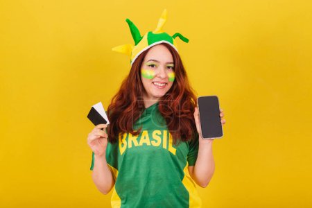 Photo for Caucasian redhead woman brazil soccer fan holding credit cards and cellphone, mobile sales concept, smartphone, app purchases. - Royalty Free Image