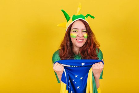Foto de Mujer caucásica pelirroja fanática del fútbol brasileño sosteniendo bandera, celebrando, vitoreando y festejando. - Imagen libre de derechos
