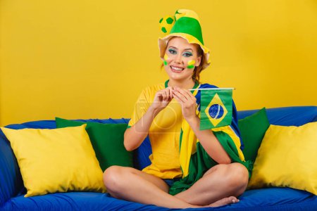 Foto de Mujer caucásica, pelirroja, aficionada al fútbol brasileño, brasileña, en un sofá con bandera de Brasil - Imagen libre de derechos