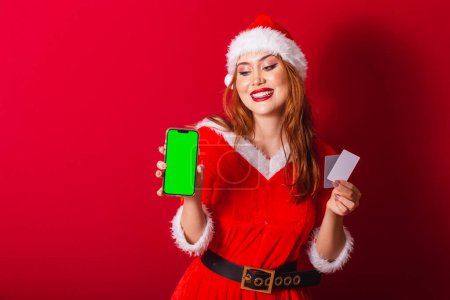 Photo for Beautiful Brazilian red-haired woman, dressed in Christmas clothes, Santa Claus. holding credit cards and smartphone with green screen in chroma. - Royalty Free Image