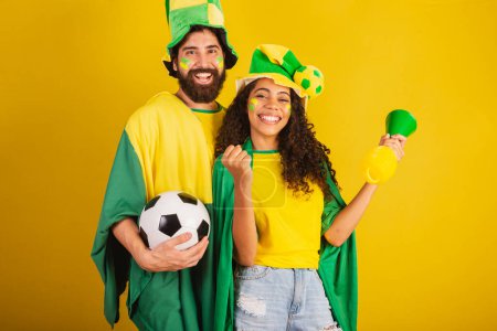 Foto de Pareja de aficionados al fútbol brasileño, vestidos con los colores de la nación, mujer negra, hombre caucásico. utilizando bandera y accesorios - Imagen libre de derechos