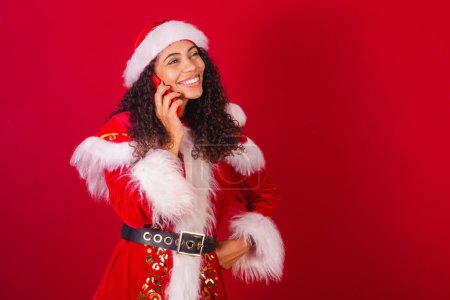 Photo for Beautiful brazilian black woman, dressed as santa claus, christmas clothes. in voice call by smartphone. - Royalty Free Image