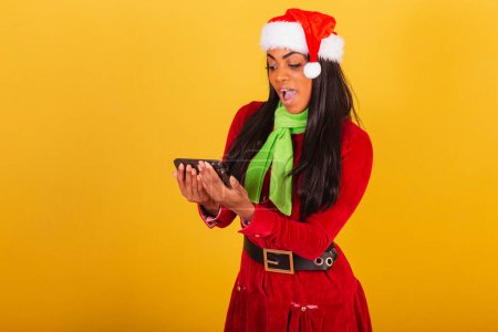 Téléchargez les photos : Belle femme brésilienne noire, vêtue de vêtements de Noël, Père Noël, smartphone et célébration. - en image libre de droit