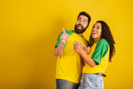 Photo for Couple of brazil soccer supporters, dressed in the colors of the nation, black woman, caucasian man. Pointing at camera. - Royalty Free Image