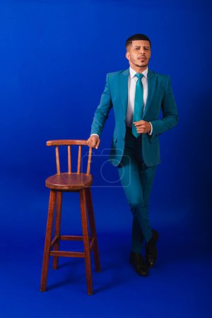 Foto de Hombre negro brasileño, vestido con traje y corbata azul. hombre de negocios. silla de madera plantea - Imagen libre de derechos