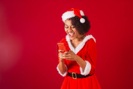 Téléchargez les photos : Belle femme brésilienne noire, habillée en Père Noël claus, maman claus, regarder quelque chose sur smartphone - en image libre de droit