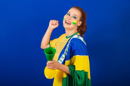 Photo for Caucasian woman, redhead, Brazil soccer fan, Brazilian, blue background, dancing with flag and horn - Royalty Free Image
