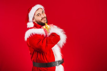 Photo for Brazilian man, dressed in santa claus clothes, embraced with red gift to camera. - Royalty Free Image